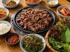 Korean BBQ table setting with grilled duck, lettuce wraps, chive salad, ssamjang, rice and side dishes on bright wooden table